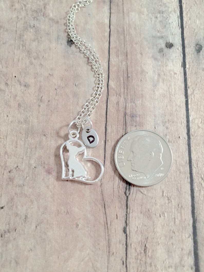 May include: Silver necklace with a heart-shaped pendant featuring a silhouette of a chihuahua. The pendant has a small charm with the letter "D" on it. The necklace is displayed next to a US dime for size comparison.