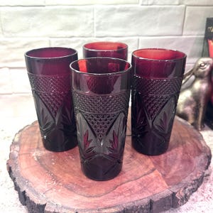 May include: Four vintage ruby red drinking glasses with diamond and leaf pattern. The glasses are arranged on a round wooden surface. The glasses are a deep red color and have a textured design.