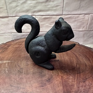 May include: A black cast iron squirrel figurine with a curved tail and outstretched paw. The squirrel is positioned on a wooden surface, showcasing its detailed features and textured finish. This decorative item adds a touch of nature-inspired charm.