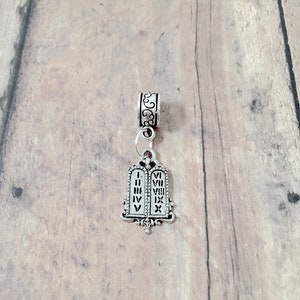May include: Silver charm featuring a pair of tablets with Roman numerals I through X, representing the Ten Commandments. The charm is attached to a silver bead with a decorative design.