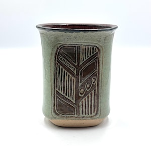 May include: A handmade ceramic tumbler with a light green exterior and a dark brown interior. The tumbler features a carved, rectangular design with a leaf-like pattern in shades of brown and tan. The rim is dark brown.