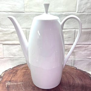 May include: A white ceramic coffee pot with a long spout and a curved handle. The pot has a lid with a small, vertical handle. The pot is sitting on a wooden surface.