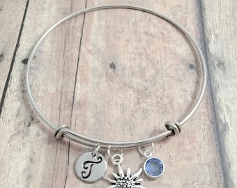 May include: Silver-toned bangle bracelet with three charms. Charms include a circular pendant with the letter "T", a sunburst design, and a small blue gemstone. The bracelet is displayed on a light brown wooden surface.