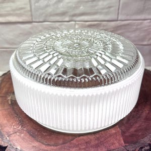 May include: A clear glass light fixture cover with a decorative, geometric pattern on top. The base is white with vertical ridges. The fixture is round and sits on a wooden surface.