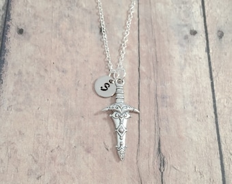 May include: Silver-toned necklace featuring a detailed dagger pendant and a small circular charm with the letter "S". The chain is delicate, and the pendant has intricate decorative elements. The necklace is displayed on a wooden surface.