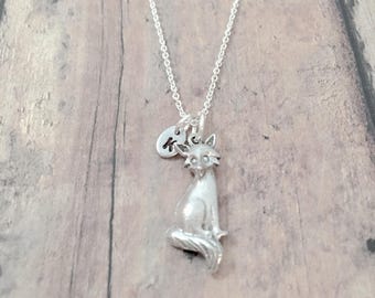 May include: A silver necklace with a small silver cat charm and a silver initial charm. The initial charm is engraved with the letter "K".