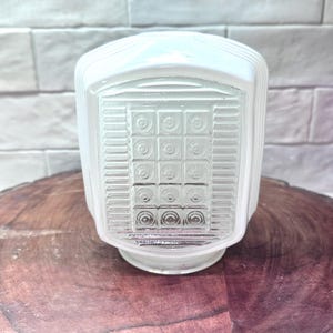 May include: A vintage, white glass light fixture with a rectangular, textured front panel featuring a grid of squares and circles. The fixture has a rounded top and a circular base. The light fixture is sitting on a wooden surface.