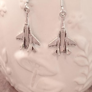 May include: A pair of silver earrings featuring a detailed jet fighter design. The earrings are on a white surface.