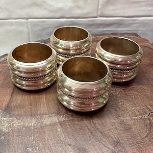 May include: Four brass napkin rings with a ribbed design and a twisted rope detail. The rings are cylindrical and sit on a wooden surface. The brass has a warm, golden hue.
