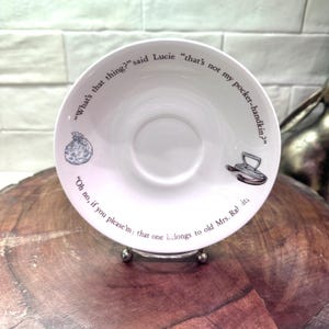 May include: White ceramic saucer with a central indentation, featuring printed text and illustrations. The saucer has a quote around the rim, with a small bag and iron illustration. The saucer is displayed on a silver stand.