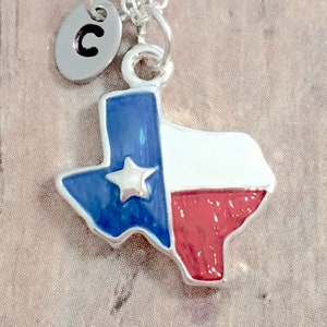 May include: A silver necklace with a charm shaped like the state of Texas. The charm is painted with the Texas flag colors: blue, white, and red. A silver charm with the letter "C" is attached to the necklace.