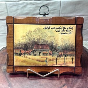 May include: A wooden framed picture with a rural landscape scene featuring a barn, windmill, and text that reads "And He will gather His wheat into the barn..." -Matthew 3:12. The frame has a metal handle and stand.
