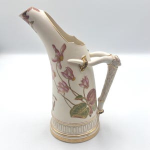 May include: A cream-colored ceramic pitcher featuring a tall, curved spout and a handle shaped like a textured antler. The body is decorated with painted pink cyclamen flowers and green leaves, accented with gold trim along the base and rim.