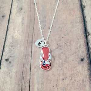 May include: A silver chain necklace with a red flip-flop charm featuring a blue and white Texas state outline. A small silver disc charm with the letter "K" is also attached to the chain.