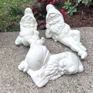 May include: Three white ceramic garden gnomes in various relaxed poses. One gnome is seated, another reclines, and the third is lying down. All are unpainted, ready for customization.