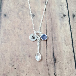 May include: A silver necklace with a charm of a small spoon, a round charm with the letter "B" and a small blue gemstone.