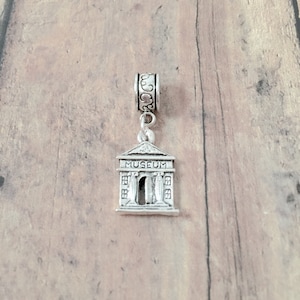 May include: Silver charm in the shape of a museum building with the word "MUSEUM" above the entrance. The charm has a decorative scroll design on the top loop.