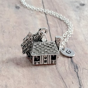 May include: A silver necklace with a charm of a small house with a tree and a chimney. The charm is attached to a silver chain with a silver initial charm that says "G".