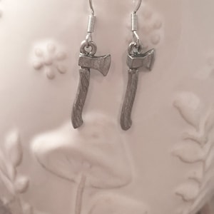 May include: A pair of silver axe earrings with a detailed design. The earrings are hanging from a white ceramic surface.