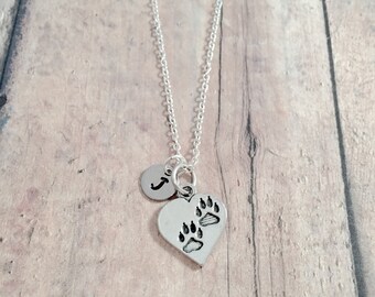 May include: A silver necklace with a heart charm featuring paw prints and a small initial charm. The necklace is on a wooden background.