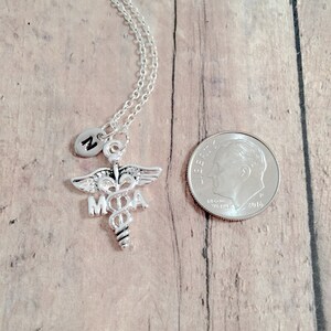 May include: Silver necklace with a medical caduceus charm and a small charm with the letter M. The necklace is displayed on a wooden surface next to a US dime.