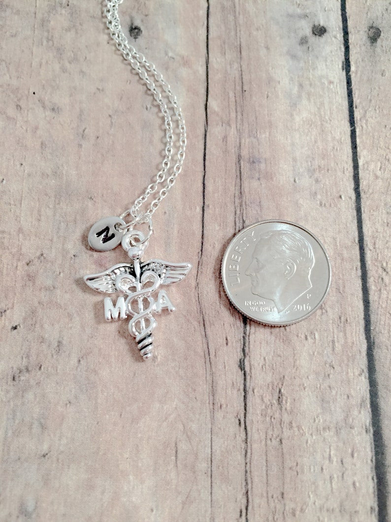 May include: Silver necklace with a medical caduceus charm and a small charm with the letter M. The necklace is displayed on a wooden surface next to a US dime.