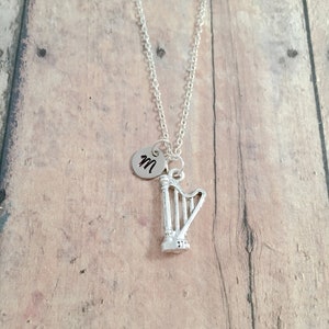 May include: A silver necklace with a small harp charm and a round charm with the letter "M" on it. The necklace is on a wooden background.