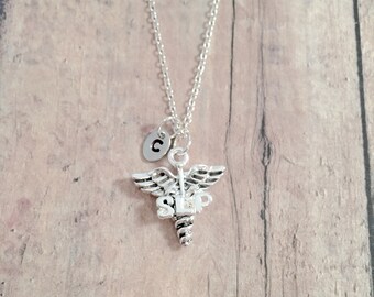 May include: Silver-toned necklace featuring a Caduceus charm, a symbol of medicine, with the letters "SLP" and a small initial pendant. The chain and charms are displayed against a light brown wooden background.