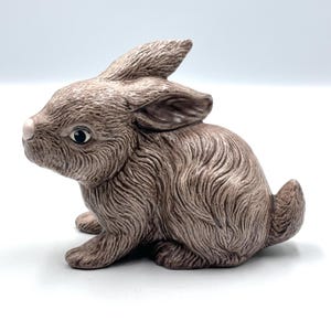 May include: A ceramic bunny figurine in shades of brown. The bunny is in a seated position with its ears perked up. It has a pink nose and detailed fur texture. The figurine is likely a decorative item, possibly for Easter or spring.