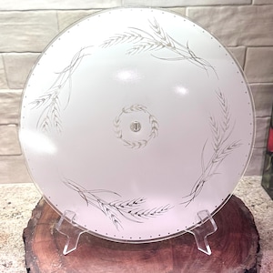 May include: A round, frosted glass ceiling light fixture with a wheat stalk design. The light fixture is supported by a clear plastic stand and is displayed on a wooden surface.