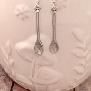 May include: Silver-toned spoon earrings with a simple design. The earrings hang from silver hooks with a coiled detail. The spoons have a rounded bowl shape. The background is a white ceramic container with floral relief.