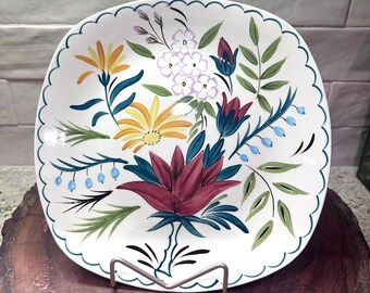 May include: A white, square ceramic plate with a scalloped edge, adorned with a vibrant floral design. The hand-painted flowers include yellow, red, blue, and purple blooms with green leaves. The plate is displayed on a metal stand.