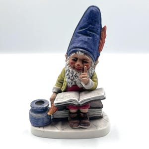 May include: A ceramic gnome figurine with a blue hat and white beard. The gnome is seated, reading a book, with an inkwell and quill nearby. The gnome is wearing a yellow jacket and red pants. The figurine is on a white base.