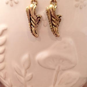May include: A pair of gold-colored earrings shaped like miniature running shoes. The earrings are suspended from gold-colored hooks. The background is a light pink ceramic container with embossed floral and mushroom designs.