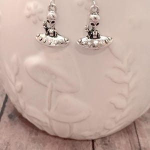 May include: Silver dangle earrings featuring a small alien figurine in a flying saucer. The alien has red eyes and is wearing a spacesuit.