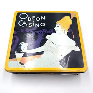 May include: Vintage metal box with a yellow border and the words "ODEON CASINO" in white. The box features an illustration of a woman in a white dress holding a cocktail. The background is black and purple.