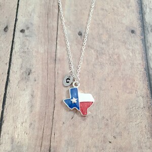 May include: Silver necklace with a charm shaped like the state of Texas, featuring the Texas flag colors of blue, white, and red. The charm also has a small silver star. The necklace has a silver initial charm with the letter "C" on it.