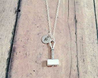 May include: A silver necklace with a small silver hammer charm and a silver initial charm. The necklace is on a wooden background.