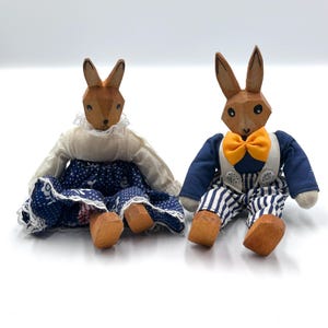 May include: Two wooden rabbit dolls. One doll wears a white blouse with a lace collar and a blue polka dot skirt. The other doll wears a blue jacket, striped pants, and an orange bow tie. Both dolls have carved wooden heads and feet.