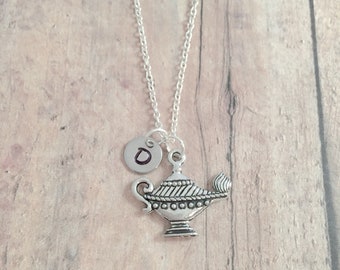 May include: Silver-toned necklace featuring a genie lamp pendant and a small circular charm with the letter "D". The chain is delicate, and the lamp pendant has intricate detailing. The necklace is displayed against a light brown wooden background.