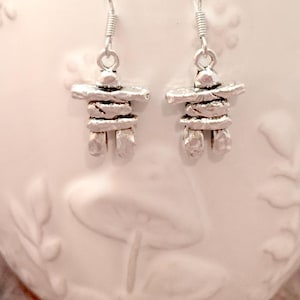 May include: Silver earrings featuring a small inukshuk charm. The inukshuk is a traditional Inuit stone structure.