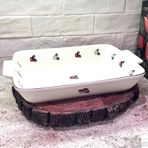 May include: A rectangular, white ceramic baking dish with small floral patterns in red, white, and green. The dish has a burgundy trim and handles on each end. It rests on a round, dark wooden base, suitable for serving.