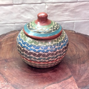 May include: A round ceramic jar with a matching lid, featuring a detailed geometric pattern of interlocking diamonds in shades of blue, green, red, and dark brown. The surface has a matte finish with visible etched lines separating each colored shape.
