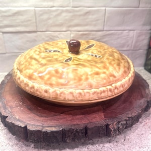 May include: A ceramic pie dish with a lid, featuring a brown crust and a decorative apple-shaped knob. The dish is placed on a round, dark wooden serving board.