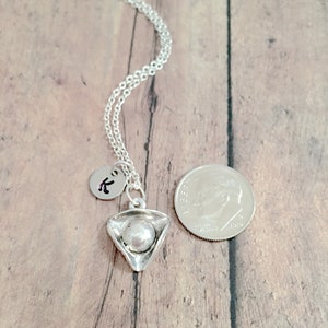 May include: A silver necklace with a small silver charm shaped like a pizza slice. The charm has a pearl-like bead in the center. The necklace also has a small silver charm with the letter "K" on it. The necklace is shown next to a US dime for size comparison.