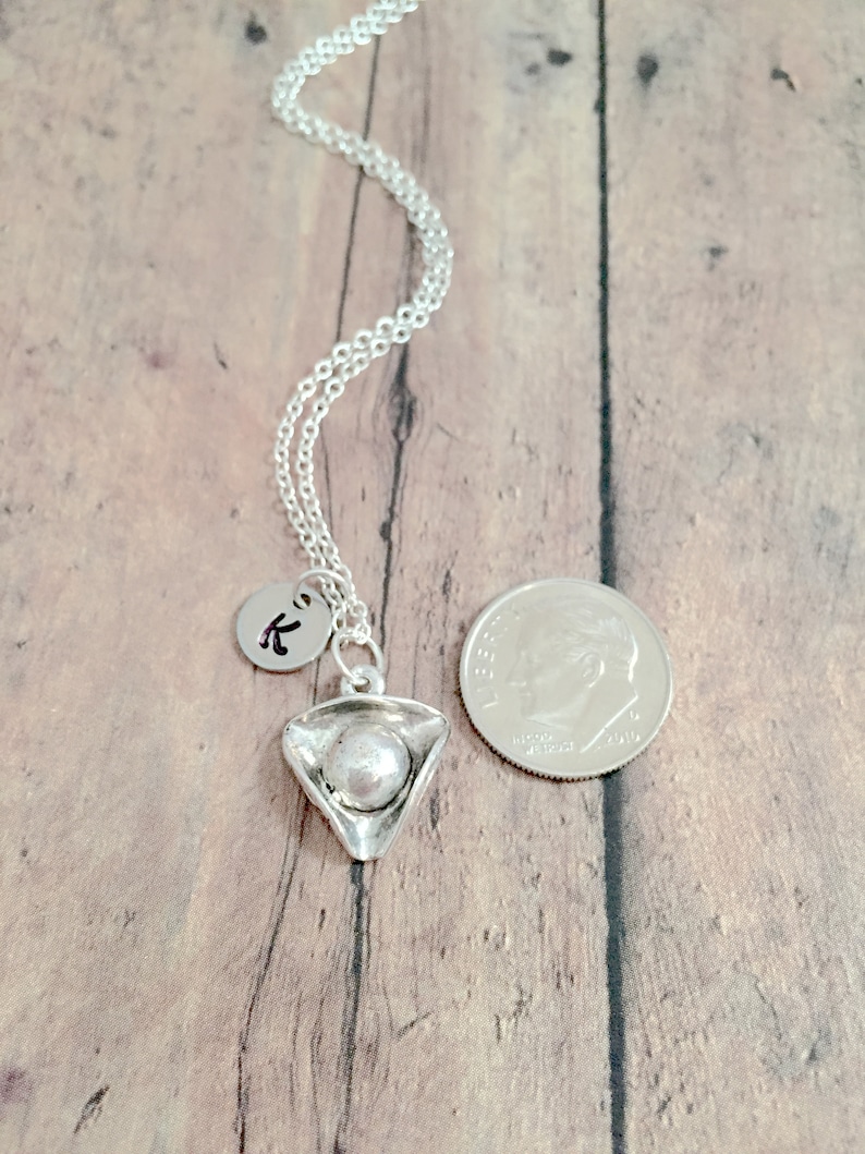 May include: A silver necklace with a small silver charm shaped like a pizza slice. The charm has a pearl-like bead in the center. The necklace also has a small silver charm with the letter "K" on it. The necklace is shown next to a US dime for size comparison.