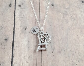 May include: A silver necklace with a spinning wheel charm and a personalized initial charm. The initial charm is a circle with the letter "M" engraved on it.