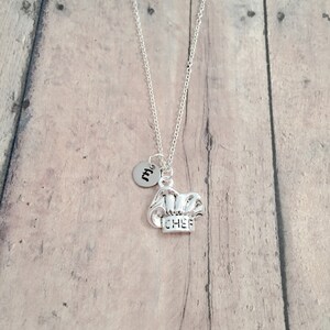 May include: Silver necklace with a chef hat charm and a small round charm with the letter "M" on it. The necklace is on a wooden background.