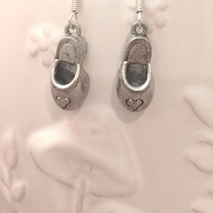 May include: A pair of silver-toned earrings featuring small shoe charms. Each charm has a heart detail and a curved top. The earrings hang from silver-colored fish hook ear wires.