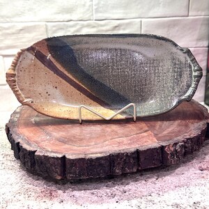 May include: A handcrafted ceramic serving platter with a unique design. The platter features a blend of colors, including brown, tan, and blue, with a textured surface. It is displayed on a wooden base and supported by a metal stand.